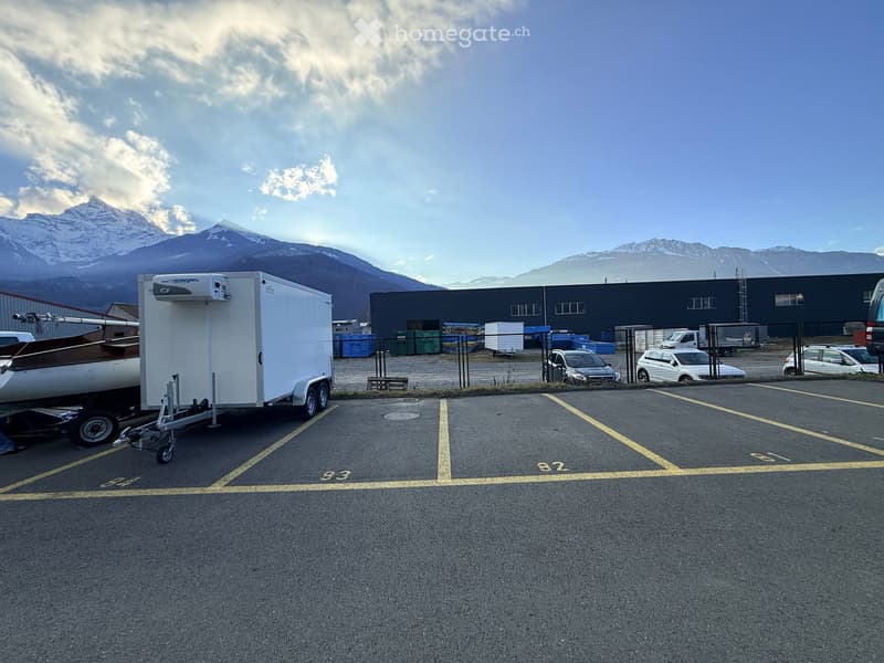 Empty parking lot with yellow lines, mountains in the background, a white trailer with a boat, and a building with a ladder.