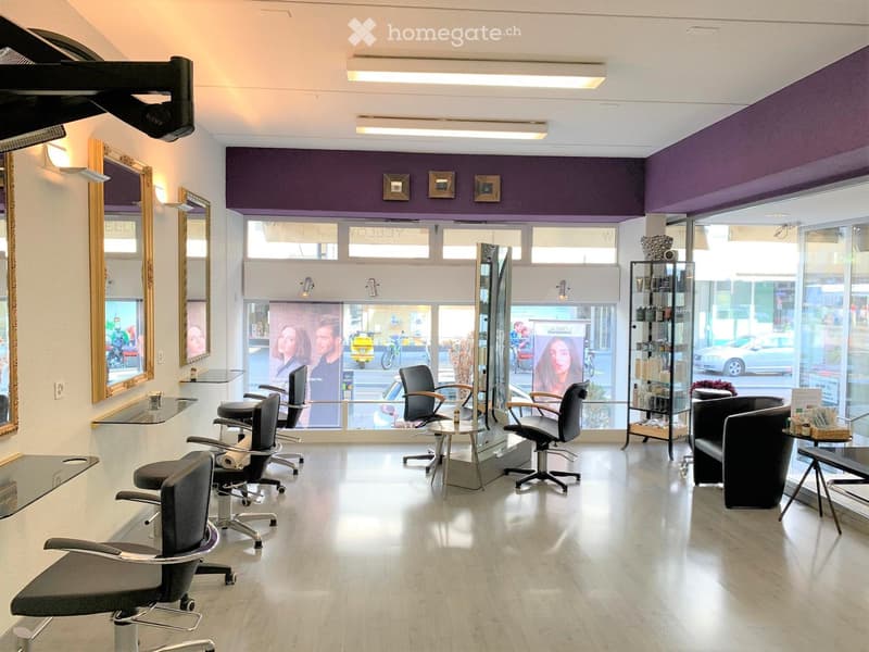 The room is well-lit with bright lights mounted on the ceiling. It has a modern and clean design with a glossy floor. There are several black swivel chairs and mirrors mounted on the wall. There are tables and shelves with bottles and other items on them.