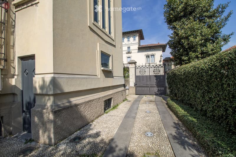 Facade of a building with a driveway leading to a closed gate, surrounded by hedges and cobblestones