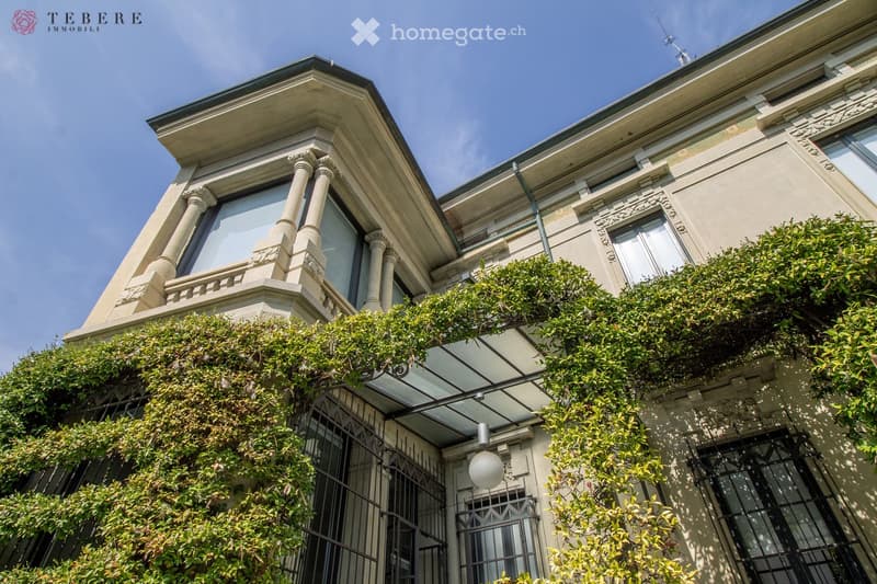 Facade of a large house with a balcony, covered by climbing plants, large windows, and a lamp