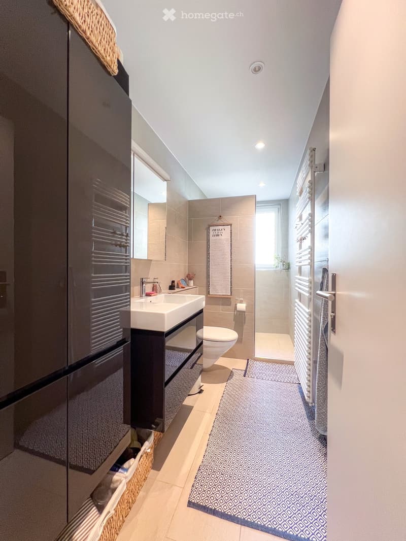 Bathroom with white sink, black cabinet, white toilet, shower, blue and white rug, white tiles, mirror, towel rack