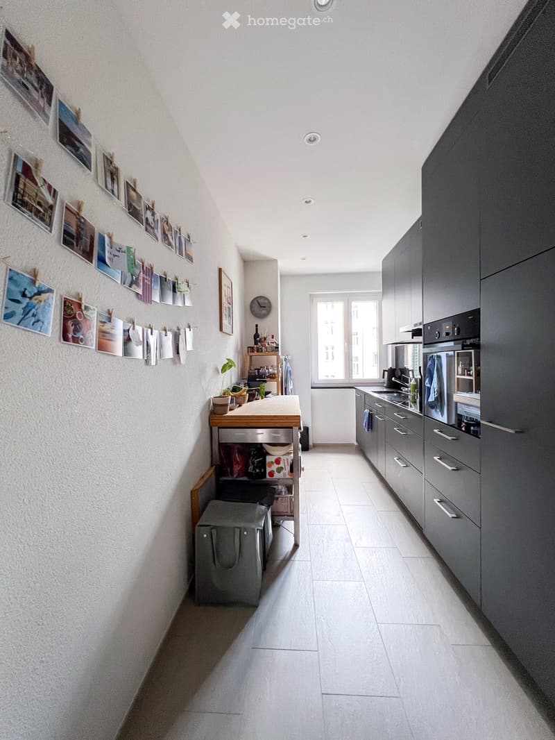 modern kitchen with white walls, wooden table, gray cabinets, white tiled floor, window, plants, clock, and pictures on the wall