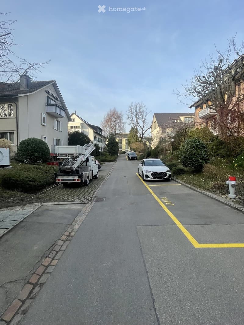 A street with parked cars, a truck, and a fire hydrant. The road has yellow markings and is surrounded by buildings and greenery.