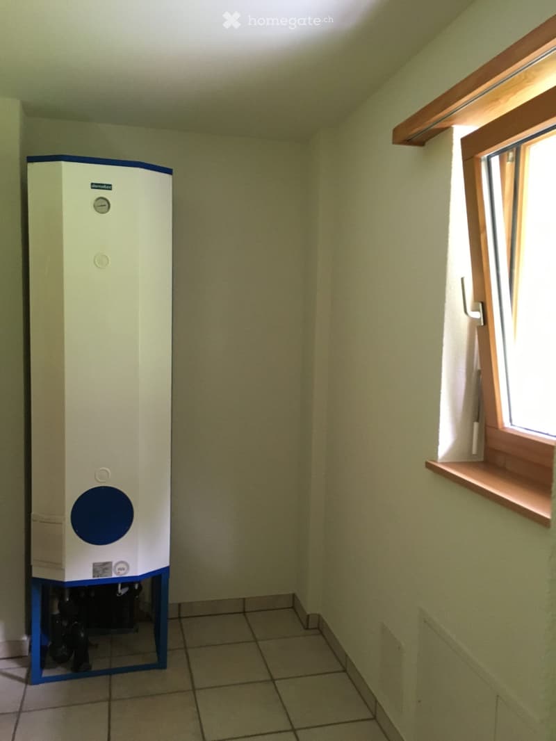 A room with a white and blue water heater, white tiled floor, white walls, and a window with a wooden frame.