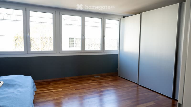 Empty room with wooden floor, large windows, gray walls, and a bed with a blue blanket.