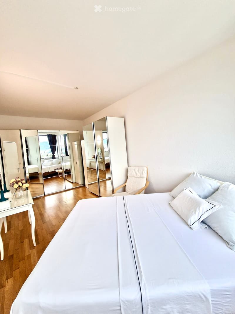 large bed, white linens, white pillows, white nightstands, white chair, mirrored closet doors, wooden floor