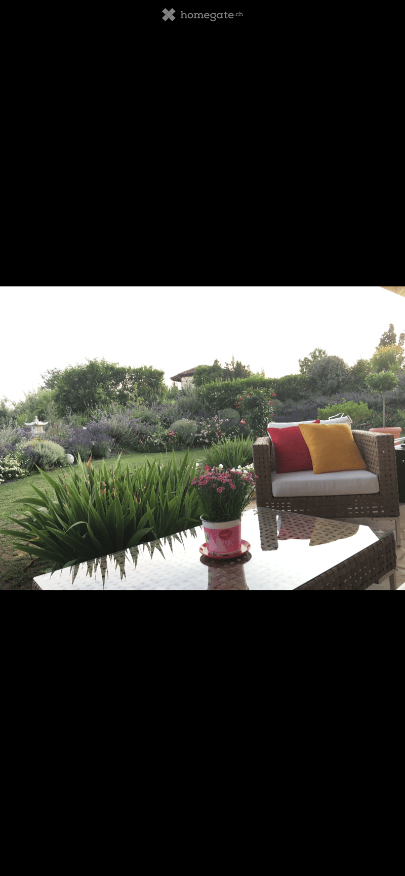 Outdoor seating area with a wicker couch, a glass table, and a flower pot. The garden has a variety of plants and flowers, including lavender, and a small white lantern. The background shows a landscape with trees and bushes.