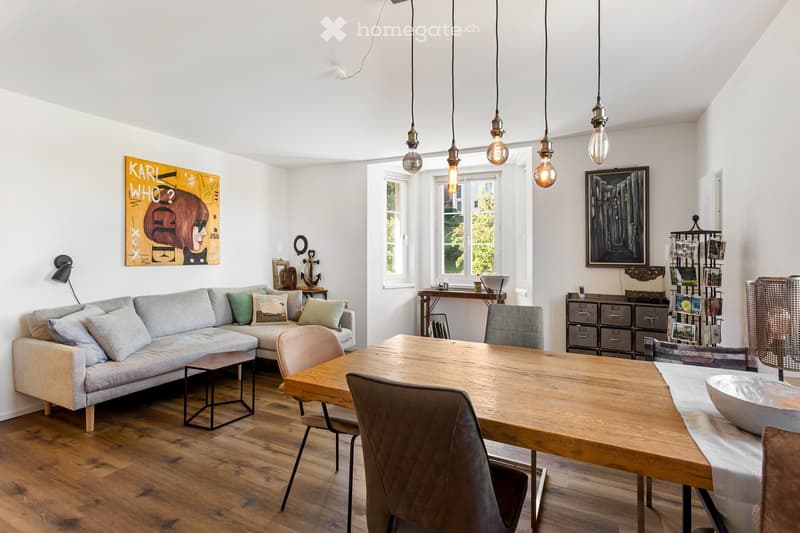 Living room with wooden floor, gray couch, wooden dining table, dining chairs, wooden side table, wall art, pendant lights, side table with bowl, cabinet with drawers, and a lamp.