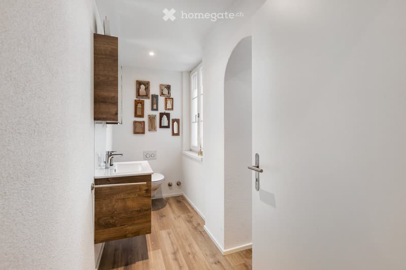 Bathroom with wooden floor, white walls, wooden cabinets, sink, toilet, window, and picture frames on the wall.