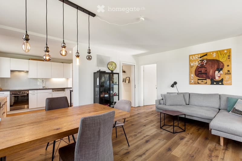 Dining area with wooden table, chairs, and a gray sofa. The room has a white wall, a painting, and a cabinet. The kitchen has white cabinets, a stove, and a sink.