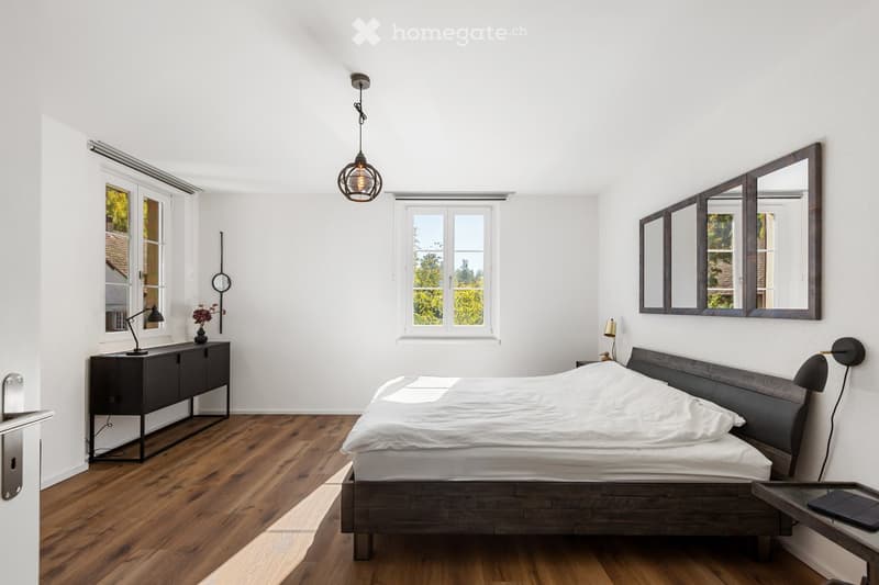 Bedroom with wooden floor, white walls, large windows, bed, nightstands, lamps, mirror, pendant light, and decorative items.