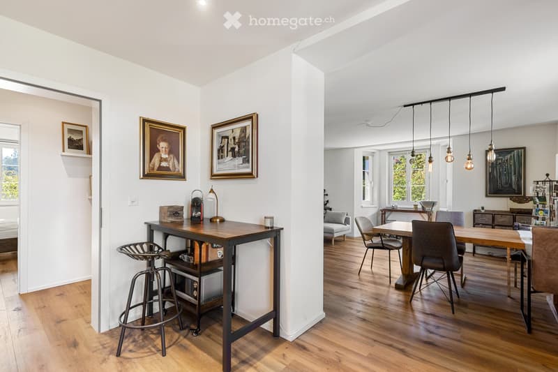 Dining area with wooden table, chairs, and a bar stool. A white wall with picture frames and a wooden floor.