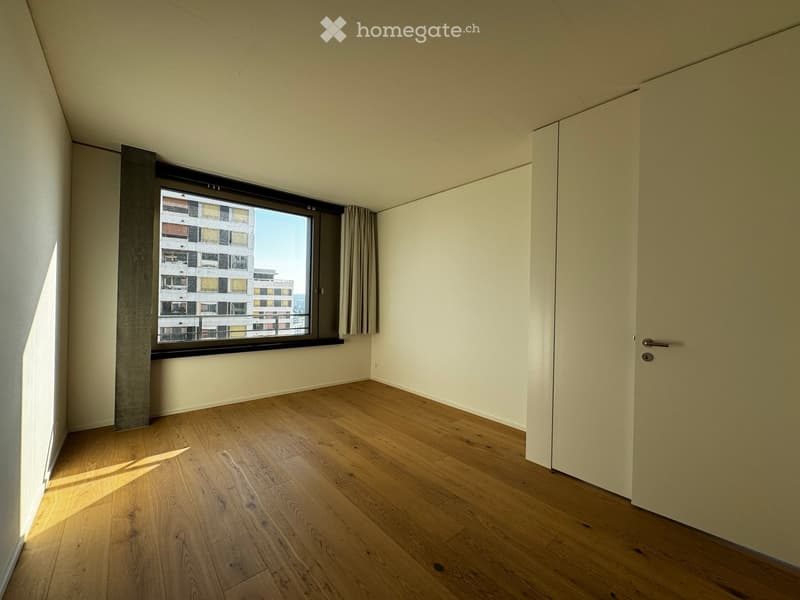 Empty room with wooden flooring, white walls, and a large window with a view of a building outside.