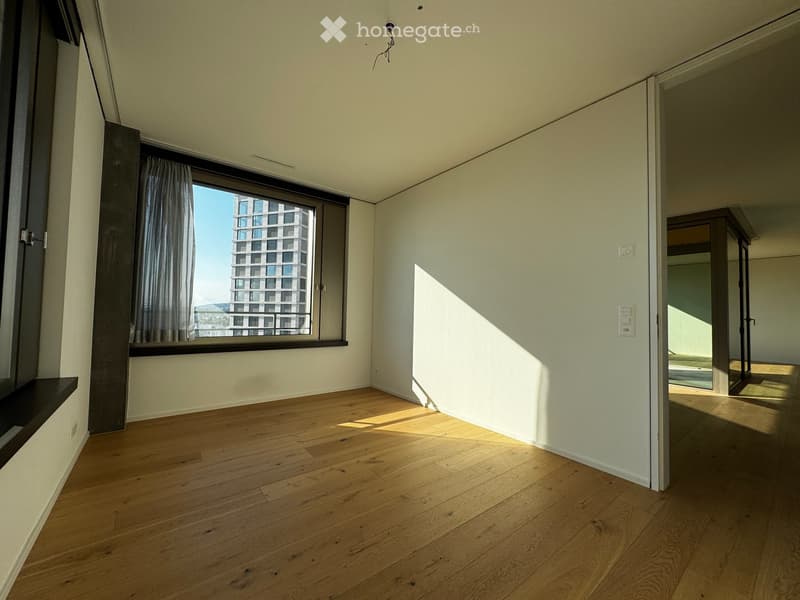 Empty room with wooden flooring, large window with curtains, and a view of a modern building outside.