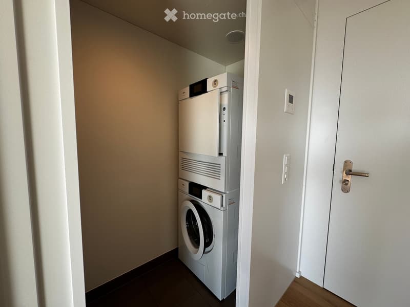 A small, well-lit room with a white washing machine and a white dishwasher stacked vertically, with a closed white door on the right side.