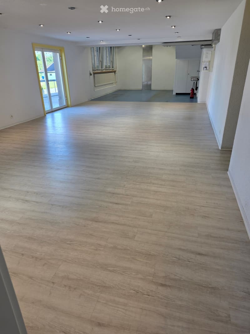 Empty room with light wooden flooring, white walls, and recessed ceiling lights. Features a large sliding glass door leading to an outdoor area and a small adjacent room with a radiator and window.