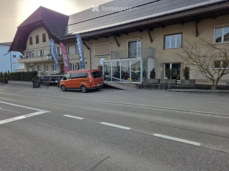 A two-story building with solar panels on the roof, a glass-enclosed entrance, and bicycles parked outside. The building has multiple windows and a balcony on the second floor. A red van is parked in front of the building.