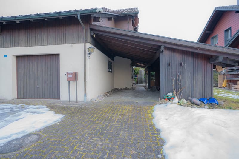 House with a garage, wooden exterior, brick wall, covered driveway, snow on the ground