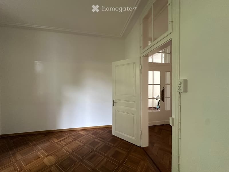 Empty room with wooden parquet flooring, white walls, and a door leading to another room with a bicycle visible in the background.