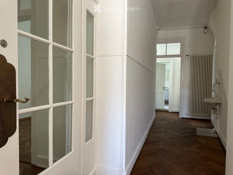 A narrow hallway with wooden flooring, white walls, and a partially visible bathroom area at the end. The hallway features a door with glass panels on the left and a radiator on the right wall.