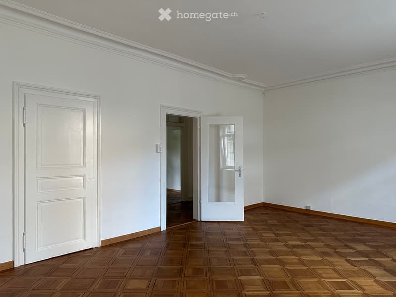 Empty room with wooden parquet flooring, white walls, and a white door. A small, mirrored glass door leads to another room.