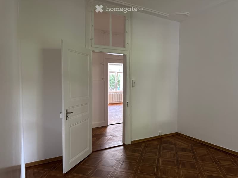 Empty hallway with wooden flooring, white walls, and an open door leading to another room with a view of a balcony area.