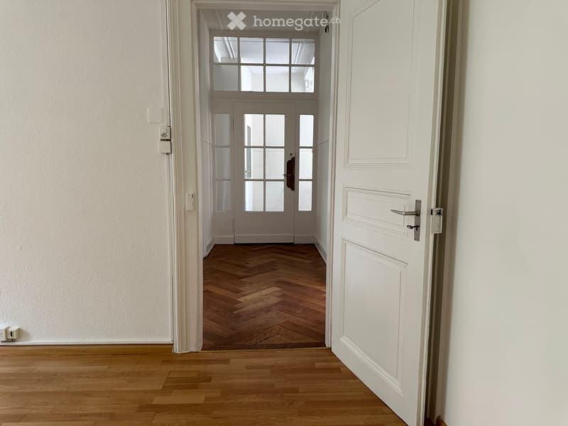 Empty hallway with wooden flooring and white walls, featuring an open doorway leading to another room with glass-paneled doors and a window above.