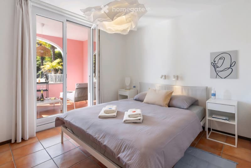 double bed, white walls, white tiled floor, glass door leading to balcony, white nightstands, decorative painting, ceiling lamp