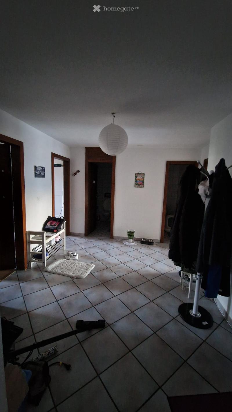 Empty room with tiled floor, white walls, wooden doors, small table with bag, hanging lamp, clothes on rack, bowl, shoes, and other items