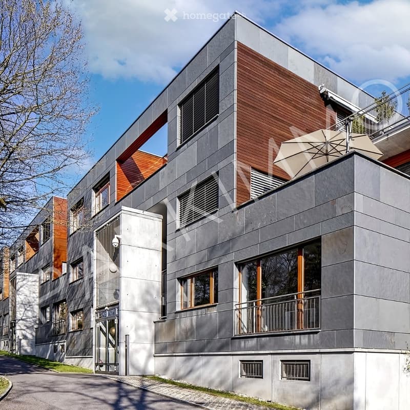 Modern multi-story building with grey and brown facade, large windows, and a rooftop terrace with an umbrella.