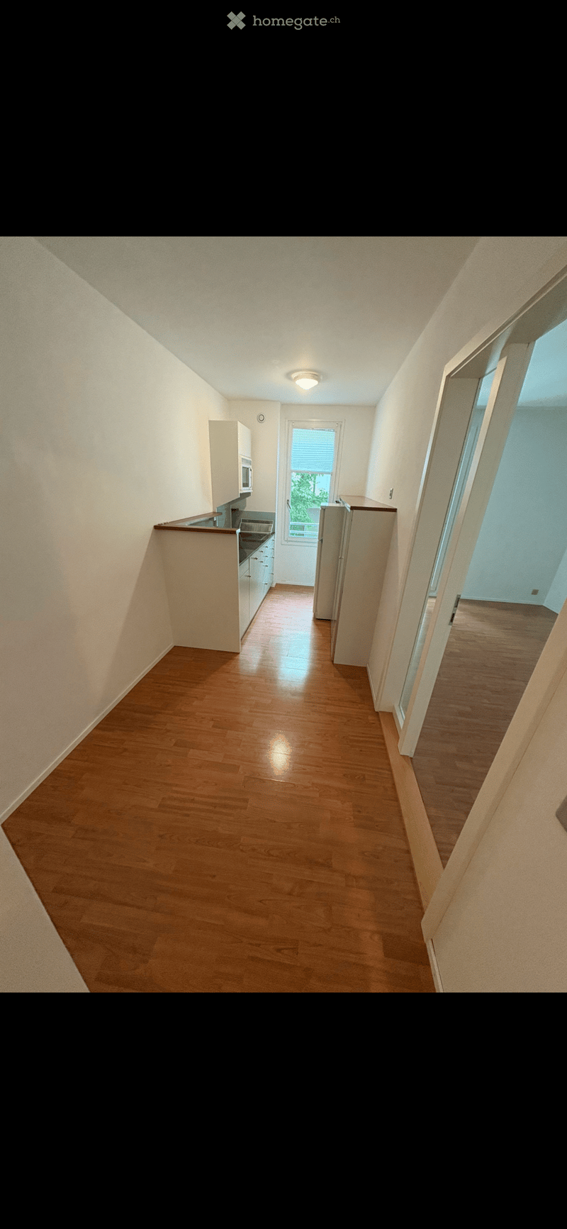 Empty room with wooden floor, white walls, kitchen area with sink and microwave, window with blinds, and a door leading to another room
