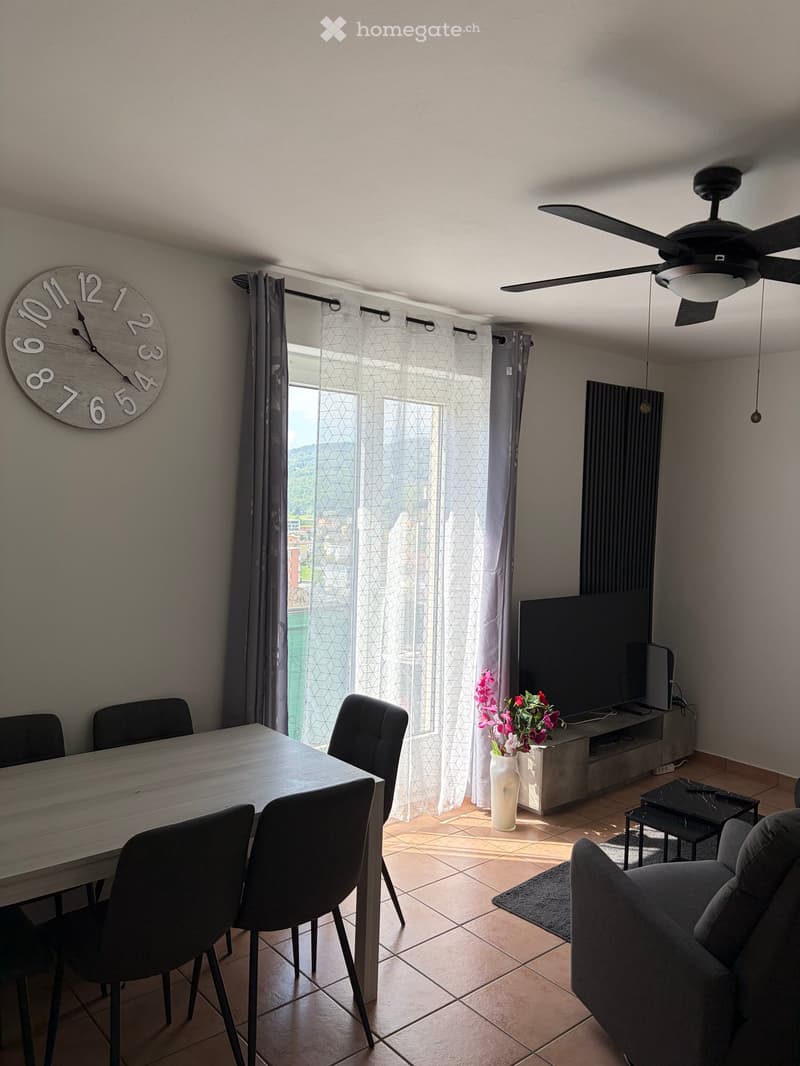 Dining area with a table and chairs, a clock on the wall, a television, a vase with flowers, and a couch