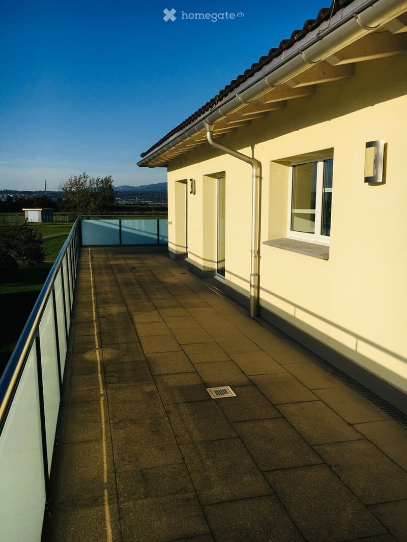 Balcony with glass railing, tiled floor, metal pipe on wall, two wall lamps, window with white frame