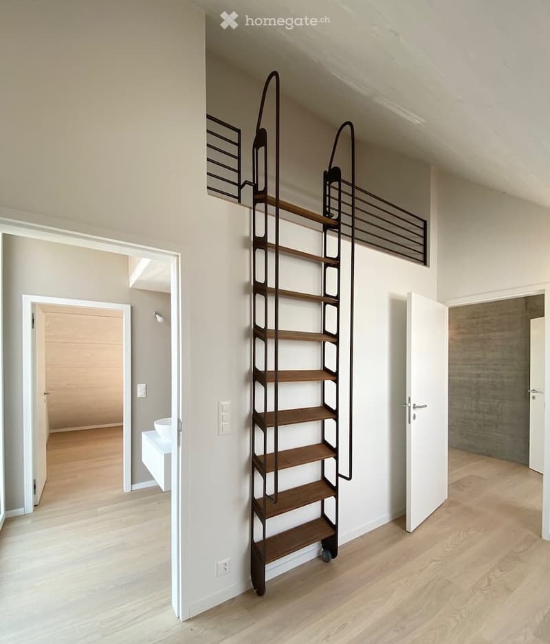 A modern hallway with a sleek, black metal staircase featuring wooden steps, leading to an upper level. The hallway has light-colored walls, wooden flooring, and several open doors leading to other rooms.