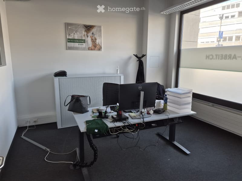 An office with a desk, computer monitor, keyboard, mouse, and various office supplies. There is a bag, a bottle, and a stack of folders on the desk. A radiator is mounted on the wall, and a poster is attached to the wall. A glass window is on the right si