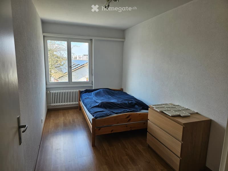 Single bedroom with wooden bed, white radiator, wooden floor, wooden nightstand, and a window with a view of trees and buildings.
