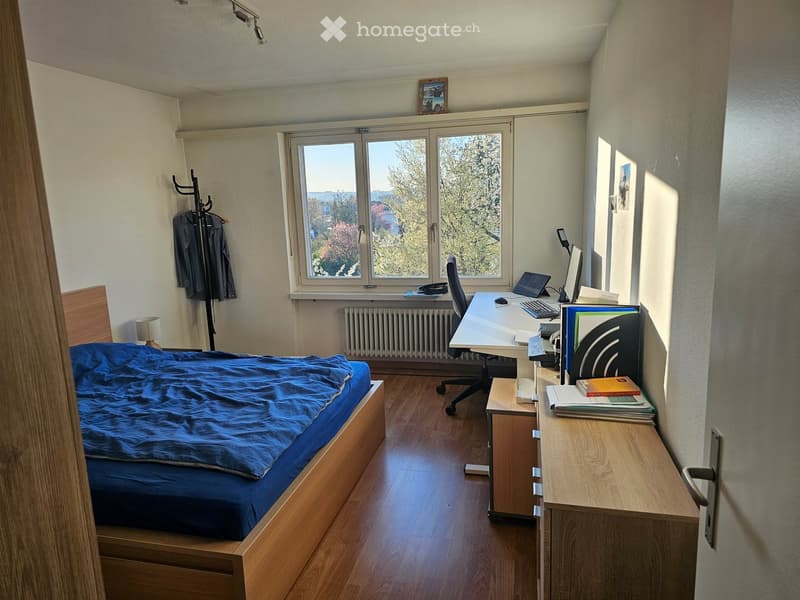 Bedroom with bed, desk, chair, radiator, window, books, picture frame, clothes on hanger, lamp, and ceiling lights.