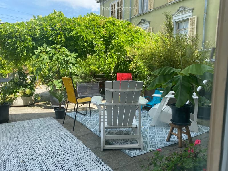 Outdoor seating area with chairs and a table, surrounded by potted plants and greenery, with a view of a building in the background.