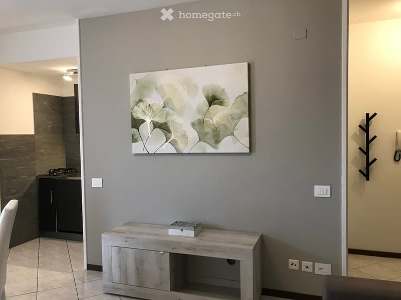 A room with a gray wall, a wooden cabinet, a white chair, and a picture frame on the wall. The room has a tiled floor and a door on the right side. The cabinet has a drawer and a storage box on top. The picture frame has a green leaf design. The room has