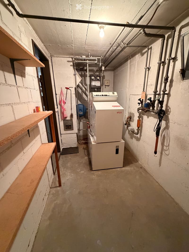 A basement room with white walls, concrete floor, wooden shelves, and two white appliances, one labeled 'elco' and the other a dishwasher.