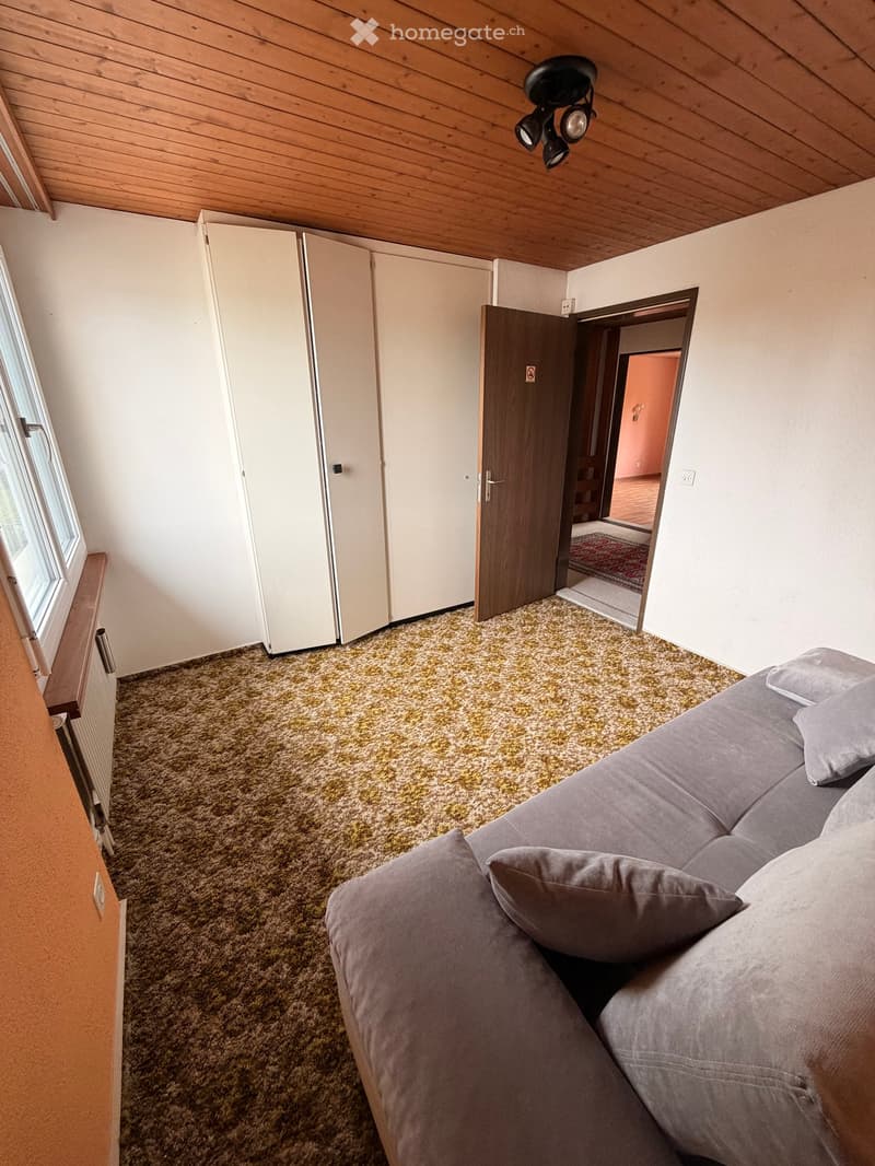 A room with a couch, carpeted floor, white walls, wooden ceiling, and a door leading to another room.