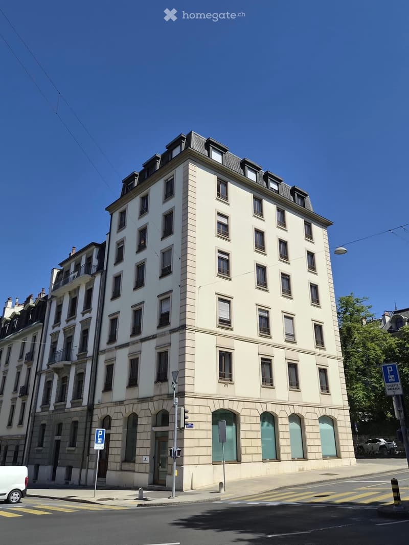 A multi-story building with light-colored facade, multiple windows, and a classic architectural style. The ground floor features arched doorways and a small entrance area.