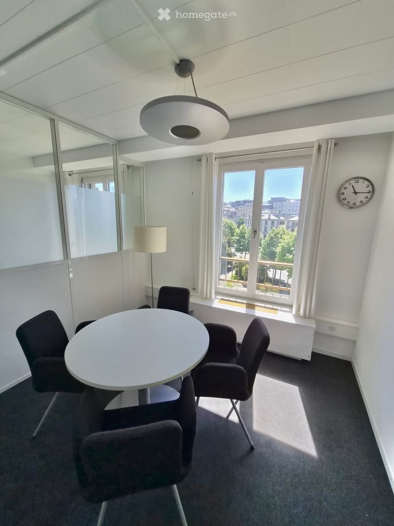 Modern furnished room with a round white table, four dark chairs, a floor lamp, a wall clock, and large windows with a view of the city.