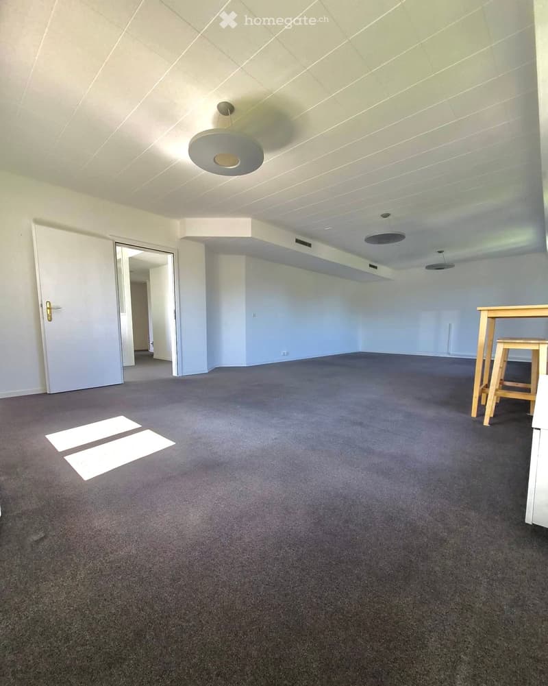 Empty room with grey carpet, white walls, and a ceiling light fixture. A door is visible on the left, and a small table and chair are placed in the corner on the right.