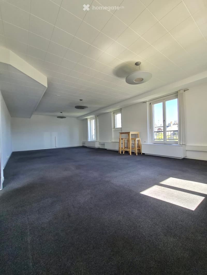 Empty room with grey carpet, white walls, large windows, and a small wooden table with two chairs in the corner.