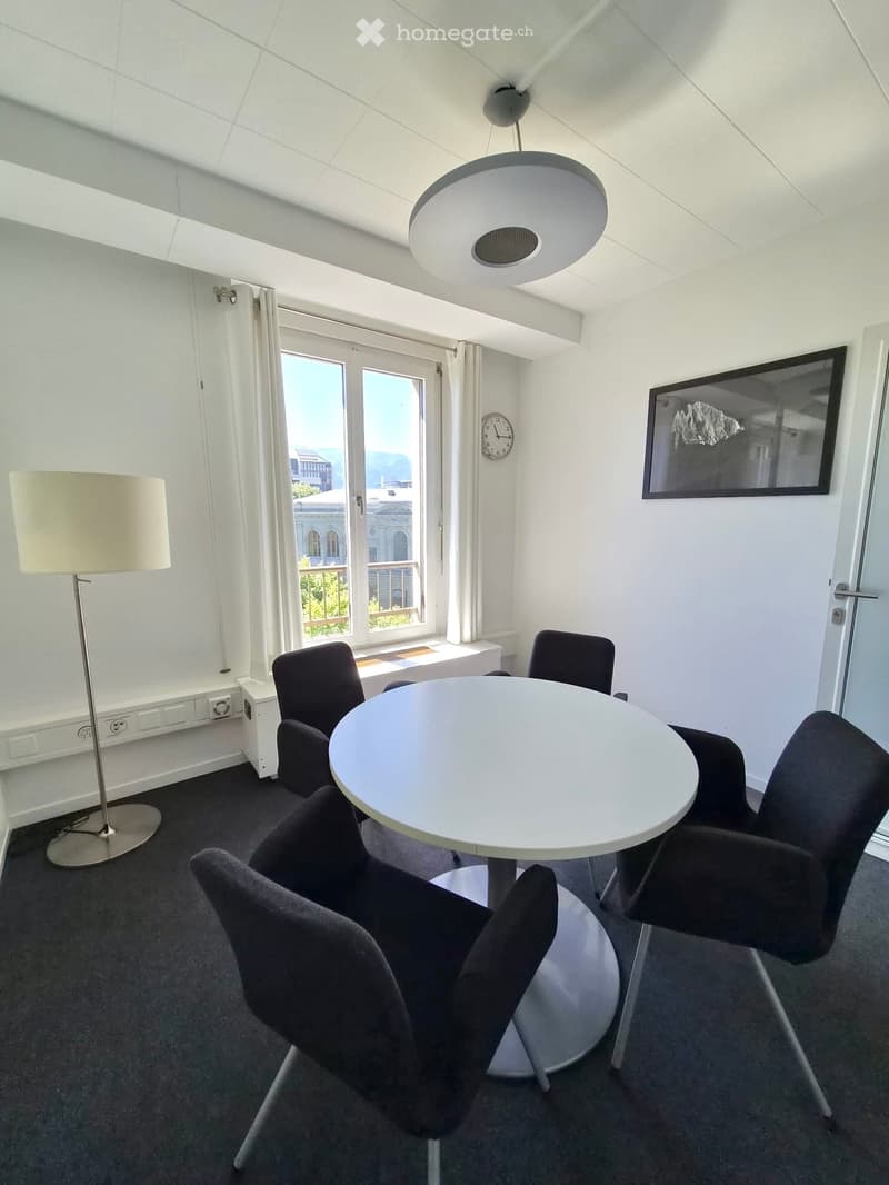 Modern, well-lit room with a round white table surrounded by black chairs, a floor lamp, a clock on the wall, and a large window with a view.