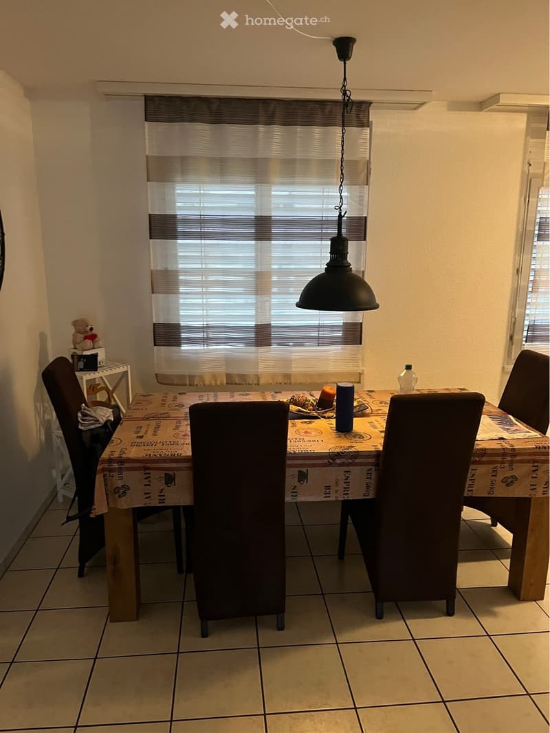 Dining room with a wooden table, four chairs, a lamp, a window with blinds, a shelf with a teddy bear, and a bottle on the table.