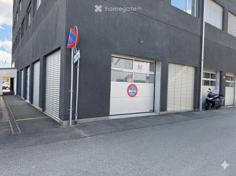 Gray building with multiple garage doors, a scooter parked in front, and a sign indicating parking restrictions.
