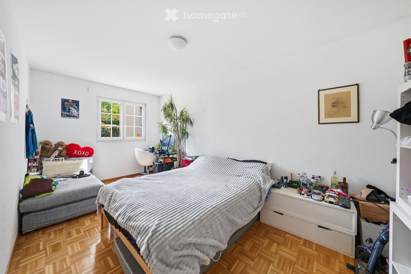 Bedroom with bed, desk, chair, plant, window, and various items on the floor and shelves.