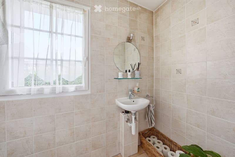Bathroom with beige tiles, white sink, mirror, window with white curtains, basket with toilet paper, plant